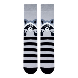 May include: A pair of grey and black striped socks with a raccoon face design. The raccoon is peeking out from behind the stripes and has a black nose and eyes.