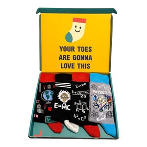 May include: A gift box containing four pairs of socks with science-themed graphics. The yellow interior lid features a cartoon sock and the text "YOUR TOES ARE GONNA LOVE THIS". The socks feature designs including a globe, Albert Einstein, and physics equations.