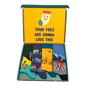 May include: A gift box containing four pairs of crew socks with cycling-themed patterns. The box lid features a yellow background with a cartoon sock illustration and the text "YOUR TOES ARE GONNA LOVE THIS" in dark blue capital letters.