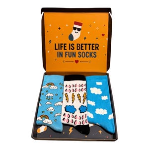 May include: A set of three pairs of socks in an open cardboard box. The box lid features the text "LIFE IS BETTER IN FUN SOCKS" with a cartoon sock graphic. The socks feature patterns of rainbows and clouds, lightning bolts and rain, and clouds.