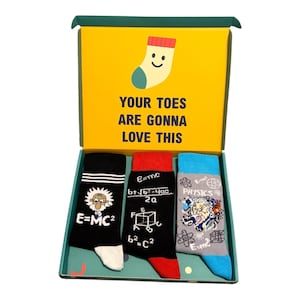 May include: A gift box containing three pairs of socks with physics-themed designs. The box lid features a yellow background with a cartoon sock and the text "YOUR TOES ARE GONNA LOVE THIS". The socks feature Albert Einstein, mathematical formulas, and physics equations in black, red, blue, white, and grey colours.