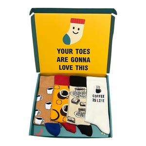 May include: A gift box containing four pairs of socks with a coffee theme. The box lid is yellow with a smiling sock graphic and the text "YOUR TOES ARE GONNA LOVE THIS." The socks feature coffee cups, coffee beans, and the phrase "COFFEE IS LIFE."