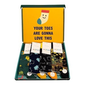 May include: A gift box containing four pairs of patterned socks with paper labels. The yellow interior lid features a cartoon sock illustration and the text: YOUR TOES ARE GONNA LOVE THIS. The socks feature green, navy, black, and camouflage designs.