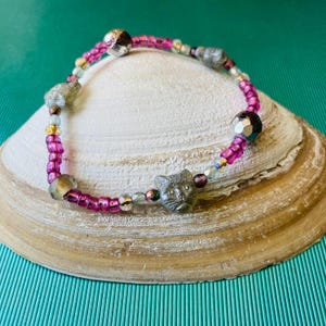 May include: A beaded bracelet featuring a cat face charm, pink beads, and silver accents. The bracelet is displayed on a seashell against a teal background. The bracelet's design incorporates various bead sizes and colors.
