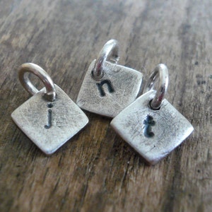 May include: Three silver charms in the shape of a square with rounded corners. Each charm has a letter stamped on it: "j", "n", and "t".
