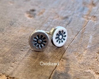 May include: A pair of silver oxidized stud earrings with a flower design. The earrings are round and have a black center with a silver flower design.