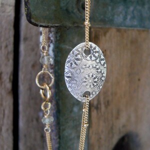 May include: A silver oval pendant with a textured surface hangs from a gold chain with small beads. The chain is double-stranded and has a small, clear crystal bead.