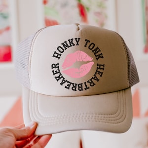 Puede incluir: Una gorra de camionero beige con un gráfico en blanco y negro que dice "Honky Tonk Heartbreaker" con una impresión de labios rosa en el centro.