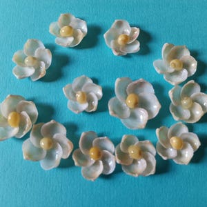 May include: A collection of light blue flower-shaped beads with yellow centers, arranged on a turquoise background. The beads have a pearlescent sheen, suggesting a delicate, handcrafted nature. These could be used for jewelry or craft projects.
