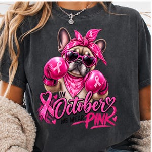 Peut inclure: Un t-shirt gris anthracite orné d'un bouledogue français portant un bandana rose, des lunettes de soleil en forme de cœur et des gants de boxe roses avec des rubans. Le motif inclut le texte "In October We Wear Pink" et un ruban rose pailleté.