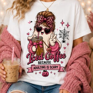 Peut inclure: Un t-shirt blanc avec un motif de fantôme portant des lunettes en forme de cœur et un nœud léopard, tenant un café glacé. Le texte indique "Iced Coffee BECAUSE ADULTING IS SCARY" avec une toile d'araignée, des cœurs roses et une citrouille.