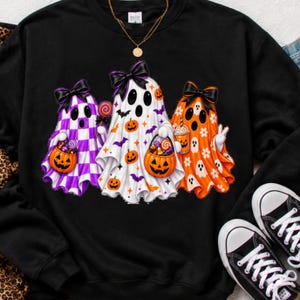 Peut inclure: Un sweat-shirt noir à col rond orné d'un motif représentant trois fantômes portant des nœuds noirs. Le fantôme de gauche a un motif à damier violet et blanc, celui du milieu est blanc avec des citrouilles orange, et celui de droite est orange.