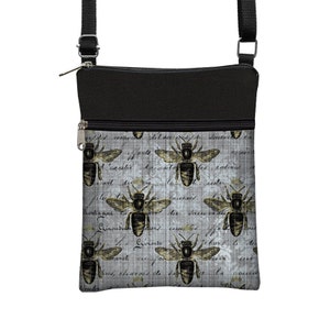 May include: A crossbody bag with a black top and strap, featuring a gray background with a repeating pattern of bees and script. The bees are detailed with brown and gold accents, and the bag has a zippered closure.