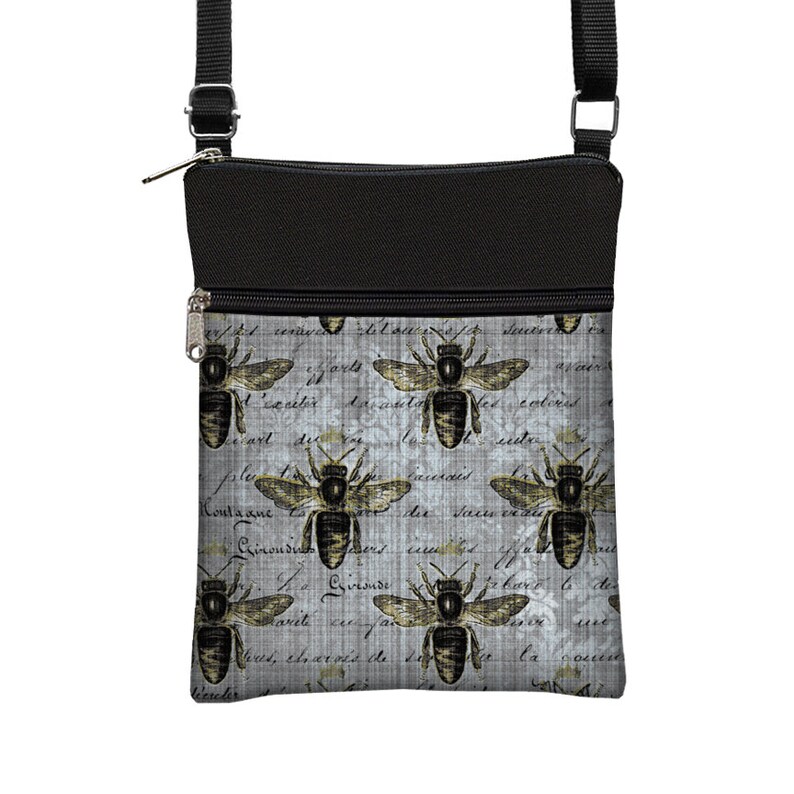 May include: A crossbody bag with a black top and strap, featuring a gray background with a repeating pattern of bees and script. The bees are detailed with brown and gold accents, and the bag has a zippered closure.