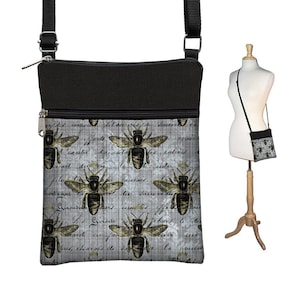 May include: A black and gray crossbody bag with a bee pattern. The bag features a black top section and a gray body with repeating bee illustrations and script. The bag has a black adjustable strap and a zipper closure.