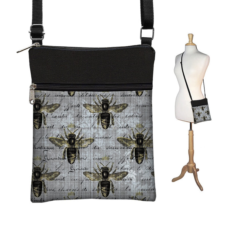 May include: A black and gray crossbody bag with a bee pattern. The bag features a black top section and a gray body with repeating bee illustrations and script. The bag has a black adjustable strap and a zipper closure.