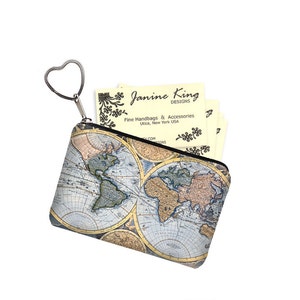 May include: A small zippered pouch with a heart-shaped keychain. The pouch is printed with a vintage world map design in shades of blue, brown, and yellow. The pouch is partially open, revealing a stack of business cards with the text "Janine King Designs Fine Handbags & Accessories Utica, New York USA" printed on them.