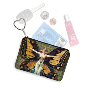 May include: A small zippered pouch with a colorful butterfly design. The pouch has a heart-shaped zipper pull and a black zipper. The butterfly is yellow, brown, and blue with a woman's face in the center. The pouch is sitting on a white surface with a credit card, coins, and a tube of lip gloss.