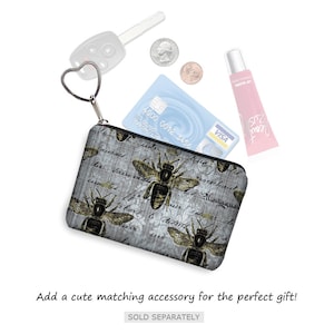May include: A small, rectangular coin purse with a zipper closure and a heart-shaped key ring. The purse features a gray background with bee illustrations and script. A credit card, coins, car key, and lip gloss are also visible.