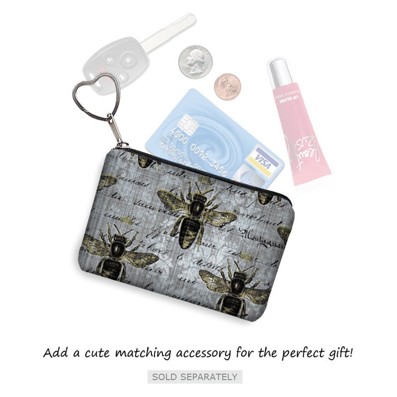 May include: A small, rectangular coin purse with a zipper closure and a heart-shaped key ring. The purse features a gray background with bee illustrations and script. A credit card, coins, car key, and lip gloss are also visible.
