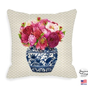 Pode incluir: Almofada decorativa quadrada com um design floral em aquarela. Apresenta um buquê de peônias rosa e vermelhas em um vaso azul e branco. O fundo tem um padrão dourado repetitivo. A almofada é rotulada como "Janine King Designs" e "Made in USA".