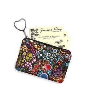 Puede incluir: Una peque&ntilde;a bolsa negra con cremallera y un llavero en forma de coraz&oacute;n. La bolsa tiene un patr&oacute;n colorido de c&iacute;rculos y puntos en tonos de rojo, naranja, amarillo, verde, azul, morado y blanco sobre un fondo negro. La bolsa est&aacute; parcialmente abierta, revelando tarjetas de visita con el texto "Janine King Designs, Fine Handbags & Accessories, Utica, New York USA".