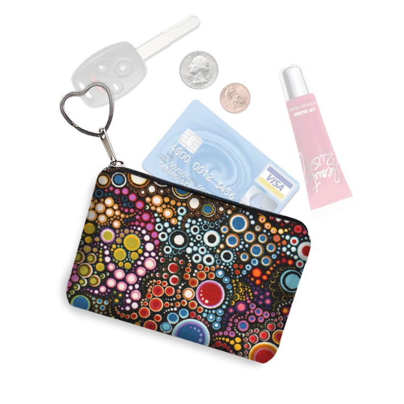 Puede incluir: Una peque&ntilde;a bolsa negra con cremallera con un patr&oacute;n abstracto colorido de c&iacute;rculos. La bolsa est&aacute; abierta y contiene una tarjeta de cr&eacute;dito con el logotipo de VISA y un b&aacute;lsamo labial rosa. Tambi&eacute;n hay una llave de coche y dos monedas sobre un fondo blanco.