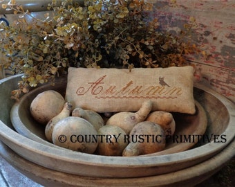 May include: A rustic wooden bowl filled with gourds and a pillow with the text "Autumn" embroidered on it. The bowl is surrounded by dried foliage.