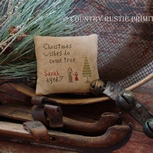 May include: A beige fabric pillow with a cross-stitch design featuring a Christmas tree and the text "Christmas wishes do come true Sarah aged 7". The pillow is surrounded by rustic wooden objects, including a metal screen, a pair of wooden ice skates, and a string of bells.