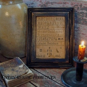 Puede incluir: Un sampler enmarcado en negro con un alfabeto vintage, números y el texto "Gracie Buckham" y "aged 12 1805". El sampler está sobre una superficie de madera con una vasija marrón y un candelabro negro con una vela encendida.