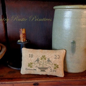 Puede incluir: Una pequeña almohada beige con un diseño de punto de cruz de dos pájaros posados ​​en una planta con tréboles de cuatro hojas. Los números 18 y 23 están cosidos a cada lado del diseño. La almohada está sentada en un estante de madera con una gran olla de barro gris y un portavelas.