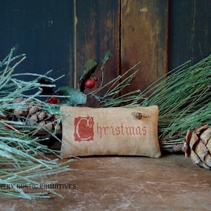 Op de afbeelding: Een rustiek bruin stoffen kussen met het woord "Christmas" geborduurd in rood. Het kussen is omringd door dennentakken en dennenappels.