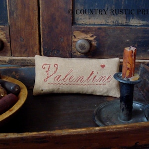 Pode incluir: Uma bandeja de madeira rústica com uma pequena almofada bordada com a palavra "Valentine" em fio vermelho. A almofada está sentada em uma bandeja de madeira com um porta-velas e uma tigela de pequenas bobinas de madeira.