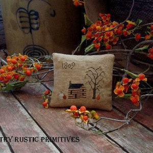 May include: A small, beige pillow with a cross-stitch design of a log cabin with a tree and the year 1869. The pillow is surrounded by a branch with orange berries.