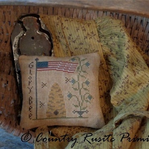 May include: A small, brown, embroidered pillow with a beehive, a plant, and an American flag. The pillow is in a wicker basket with a brown fabric.