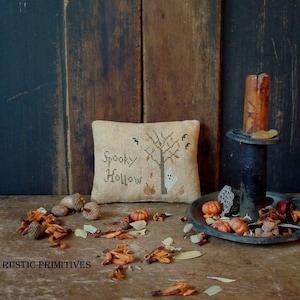 Pode incluir: Uma almofada bege com o texto "Spooky Hollow" bordado nela. A almofada apresenta um desenho de ponto cruz de uma árvore nua com um fantasma e abóboras. A almofada está sentada sobre uma superfície de madeira com decorações de outono.