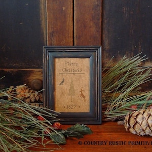 Puede incluir: Un sampler de punto de cruz enmarcado con un marco negro y un fondo de arpillera. El sampler presenta un árbol de Navidad estilizado con un pájaro, un ciervo y el texto "Merry Christmas 1827".