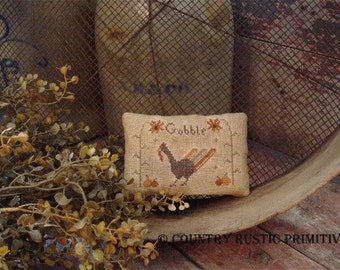 May include: A beige cushion with a cross-stitch design of a turkey with the word "Gobble" above it. The cushion is sitting on a wooden surface with a rustic metal screen behind it.