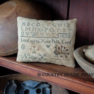 May include: A beige sampler pillow with cross-stitch embroidery. The pillow features the alphabet, a peacock, a heart, and the text "Lead me in Your Path, Lord." The pillow is signed "Nellie H." and dated "1835."