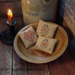 Könnte beinhalten: Drei kleine, quadratische, bestickte Kissen mit roten und schwarzen Nähten werden in einer Holzschale präsentiert. Die Kissen liegen auf einer Holzoberfläche. Die Kissen sind mit Herzen und Blumen verziert.