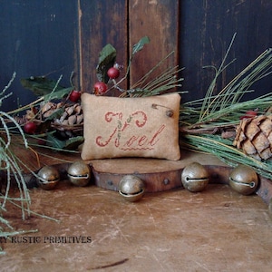 Puede incluir: Una pequeña almohada beige con bordado rojo que dice "Noel" está rodeada de ramas de pino, piñas y cascabeles.