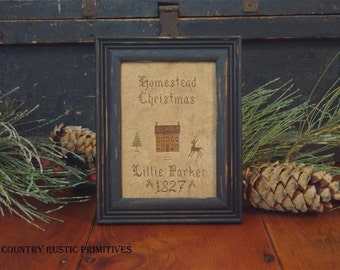 May include: A framed cross-stitch sampler with a black frame and a beige background. The sampler depicts a house, a tree, and a deer. The text reads "Homestead Christmas" and "Lillie Parker 1827".