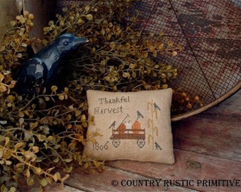 May include: A beige cushion with a cross-stitch design featuring a wagon full of pumpkins and the text "Thankful Harvest 1806". The cushion is surrounded by brown branches and a black crow.