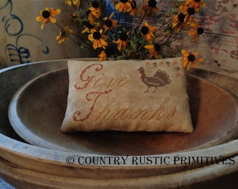 May include: A rustic brown wooden bowl with a smaller bowl inside. A cushion with the words "Give Thanks" and a stitched image of a turkey is on top of the bowls. A bouquet of yellow flowers is in the background.