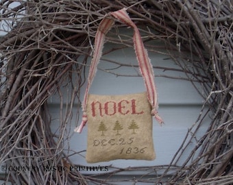 May include: A beige fabric ornament with red embroidery that reads "NOEL" and "DEC. 25 1836". The ornament is hanging from a red and white striped ribbon against a background of brown twigs.