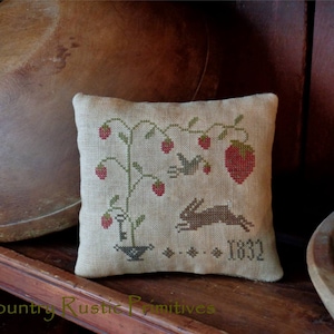 May include: A beige linen pillow with a cross-stitch design of a strawberry plant, a bird, a rabbit, and the year 1832. The pillow is sitting on a wooden shelf with a wooden bowl in the background.