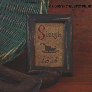 May include: A framed cross-stitch sampler with the word "Sleigh" in red lettering and a small black and red sleigh with the year "1836" below it. The frame is dark blue and the background is a natural linen fabric.