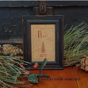 May include: A framed cross-stitch sampler with a red embroidered Christmas tree and the word "Noel" in red. The year "1875" is also embroidered in red below the tree. The frame is black and the background is a light brown.
