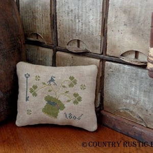 Puede incluir: Una pequeña almohada beige con un diseño de punto de cruz que presenta una maceta verde con una planta y una llave. El texto "1806" está bordado en la almohada. La almohada está sentada sobre una superficie de madera.