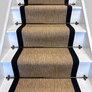 May include: A staircase featuring beige woven carpet runners with black edging. The runners are secured with metal rods and brackets on white painted stairs. The design offers a classic and elegant look for home interiors.
